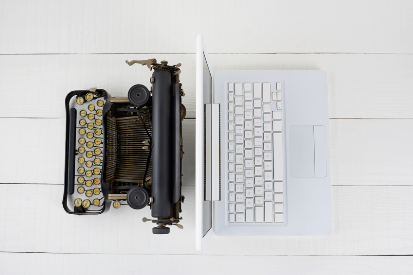 Overhead shot of and old fashioned typewriter back-to-back with a modern laptop computer on a white wood desk.